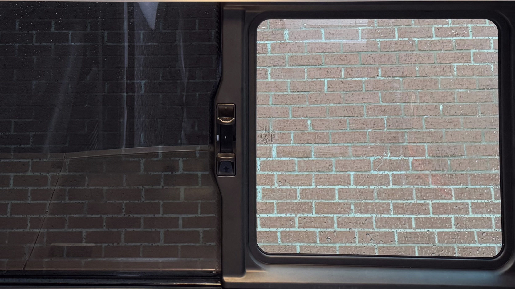 Die Fensterreihe des Busses von außen: links eine abgedunkelte Scheibe, rechts eine glasklare, durch die die Backsteinwand dahinter zu sehen ist.